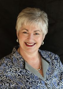 Woman in her early 60's with grey hair and a big smile.